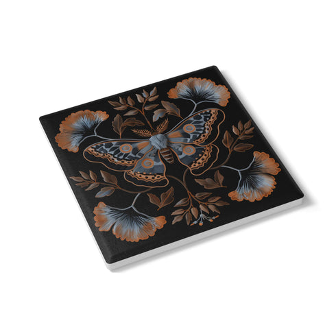 Square black ceramic coaster with detailed copper and blue moth illustration centered among symmetrical botanical elements including ginkgo-like leaves and delicate foliage.